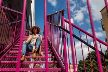 Şehirde merdivenlerde otururken şapkalı şık bir kız poz veriyor. Moda tarzı.