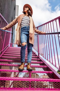 Şapkalı şık bir kız şehrin arka planına karşı korkulukların yanında poz veriyor. sokak tarzı ve moda.