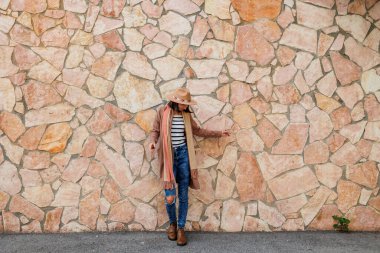 Güzel, gülümseyen bir modelin portresi. Bir şehir duvarının arkasında şapka takıyor ve şık kıyafetler giyiyor. Bir kadın sokakta bir duvarın yanında poz veriyor. Komik ve pozitif kadın dışarıda eğleniyor.. 