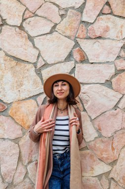 Güzel, gülümseyen bir modelin portresi. Bir şehir duvarının arkasında şapka takıyor ve şık kıyafetler giyiyor. Bir kadın sokakta bir duvarın yanında poz veriyor. Komik ve pozitif kadın dışarıda eğleniyor.. 
