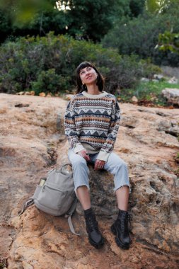 Sırt çantalı ve süveterli bir yürüyüşçü ormanda bir taşın üzerinde oturuyor.