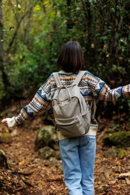 Süslü kazaklı ve sırt çantalı bir kız ormanda yürümekten zevk alıyor. Ormanda yürüyüş yapmak. Sırt çantalı bir kız ormanda yürüyor. Yürüyüş ve yürüyüş. moda ve tarz.