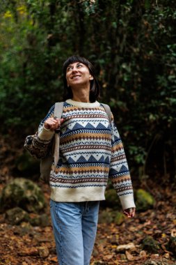 Sırt çantalı kız ormanda yürümekten hoşlanıyor. Ormanda yürüyüş yapmak. Süslü süveter giyen genç bir kız ormanda yürüyor. Yürüyüş ve yürüyüş.