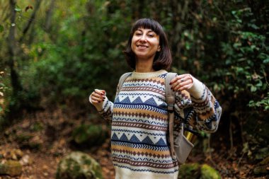 Süslü kazaklı ve sırt çantalı bir kız ormanda yürümekten zevk alıyor. Ormanda yürüyüş yapmak. Sırt çantalı bir kız ormanda yürüyor. Yürüyüş ve yürüyüş. moda ve tarz.