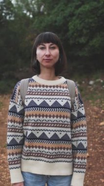 Dikey çekim. Süslü bir kazak giyen ve sırt çantalı bir kız ormanda yürümekten zevk alıyor. Yüzünüzü kapatın. Ormanda yürüyüş. Sırt çantalı bir kız ormanda yürüyor. Yürüyüş ve yürüyüş. moda ve tarz.