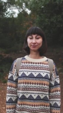 Dikey çekim. Süslü bir kazak giyen ve sırt çantalı bir kız ormanda yürümekten zevk alıyor. Yüzünüzü kapatın. Ormanda yürüyüş. Sırt çantalı bir kız ormanda yürüyor. Yürüyüş ve yürüyüş. moda ve tarz.