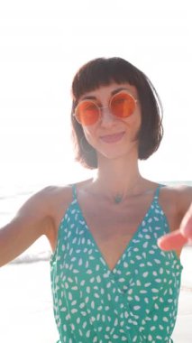 Dikey çekim. Genç kız kalp yapıyor. Aşkın sembolü. Turuncu güneş gözlüklü neşeli kadın elleriyle kalp şekli oluşturur. Kız sahilde eğleniyor..