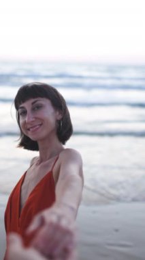 Dikey çekim. Sevgililer Günü. Sahilde elbiseli bir kız. Genç ve mutlu bir kadın adamın elini tutuyor. Beni takip edin. Elbise giymiş güzel bir kız kumsalda bir erkeğin elini tutuyor..