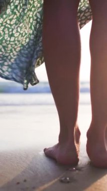 Dikey çekim. Gün batımında kumlu okyanus sahilinde çıplak ayaklı dişi bir turistin ayakları. Bir kadın yaz tatilinin tadını çıkarır. Seyahat, turizm, seyahat konsepti.
