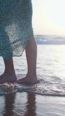 Dikey çekim. Dişi bir turistin ayakları çıplak ayakla kumlu bir okyanus sahilinde gün batımında, açık havada yürüyor. Bir kadın yaz tatilinin tadını çıkarır. Seyahat, turizm, seyahat kavramı.