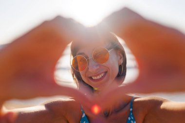 Genç kız kalp yapar. Aşkın şeklinin bir sembolü. Turuncu güneş gözlüklü neşeli kadın elleriyle kalp şekli yapıyor. Kız sahilde eğleniyor..