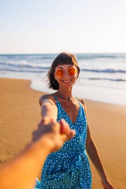 Beni takip edin. Genç ve mutlu bir kadın adamın elini tutuyor. Elbise giymiş güzel bir kız sahilde yürüyor, bir erkeğin elini tutuyor, romantik bir yürüyüş. Sahilde bir yürüyüşe çık..