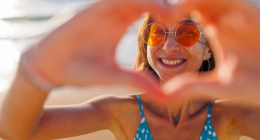 Genç kız kalp yapar. Aşkın şeklinin bir sembolü. Turuncu güneş gözlüklü neşeli kadın elleriyle kalp şekli yapıyor. Kız sahilde eğleniyor..