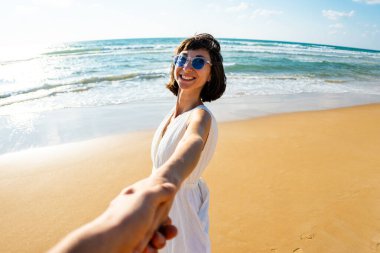 Birbirlerine aşık çiftler sahilde eğleniyorlar. Beni takip edin. Genç ve mutlu bir kadın erkeklerin elini çekiyor. Elbise giymiş güzel bir kız sahilde yürüyor, bir erkeğin elini tutuyor, romantik bir yürüyüş. Sevgililer Günü.