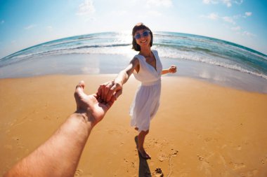 Birbirlerine aşık çiftler sahilde eğleniyorlar. Beni takip edin. Genç ve mutlu bir kadın erkeklerin elini çekiyor. Elbise giymiş güzel bir kız sahilde yürüyor, bir erkeğin elini tutuyor, romantik bir yürüyüş. Sevgililer Günü.