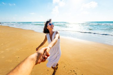 Birbirlerine aşık çiftler sahilde eğleniyorlar. Beni takip edin. Genç ve mutlu bir kadın erkeklerin elini çekiyor. Elbise giymiş güzel bir kız sahilde yürüyor, bir erkeğin elini tutuyor, romantik bir yürüyüş. Sevgililer Günü.