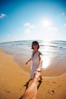 Birbirlerine aşık çiftler sahilde eğleniyorlar. Beni takip edin. Genç ve mutlu bir kadın erkeklerin elini çekiyor. Elbise giymiş güzel bir kız sahilde yürüyor, bir erkeğin elini tutuyor, romantik bir yürüyüş. Sevgililer Günü.