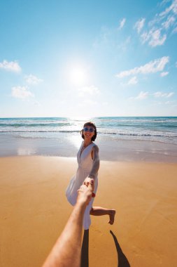 Birbirlerine aşık çiftler sahilde eğleniyorlar. Beni takip edin. Genç ve mutlu bir kadın erkeklerin elini çekiyor. Elbise giymiş güzel bir kız sahilde yürüyor, bir erkeğin elini tutuyor, romantik bir yürüyüş. Sevgililer Günü.