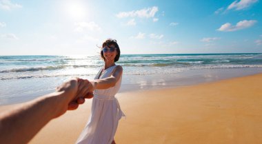 Birbirlerine aşık çiftler sahilde eğleniyorlar. Beni takip edin. Genç ve mutlu bir kadın erkeklerin elini çekiyor. Elbise giymiş güzel bir kız sahilde yürüyor, bir erkeğin elini tutuyor, romantik bir yürüyüş. Sevgililer Günü.