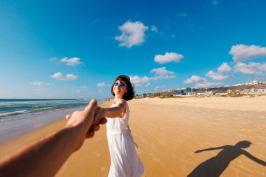 Birbirlerine aşık çiftler sahilde eğleniyorlar. Beni takip edin. Genç ve mutlu bir kadın erkeklerin elini çekiyor. Elbise giymiş güzel bir kız sahilde yürüyor, bir erkeğin elini tutuyor, romantik bir yürüyüş. Sevgililer Günü.