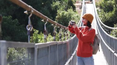 Seyahat ve fotoğrafçılık. Elinde kamera ve sırt çantası olan genç bir kadın asma köprü boyunca yürüyor. Gezinin ve fotoğraf çekmenin keyfini çıkaran mutlu bayan turist. Kameralı kız..