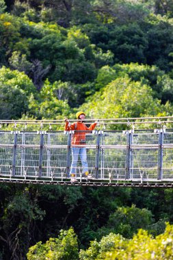 Kameralı ve sırt çantalı genç bir kadın asma köprü boyunca yürümekten hoşlanıyor. Yolculuğunun tadını çıkaran mutlu bir bayan turist. Sırt çantalı kız..