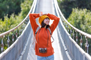 Seyahat ve fotoğrafçılık. Elinde kamera ve sırt çantası olan genç bir kadın asma köprü boyunca yürüyor. Seyahatinin tadını çıkaran mutlu bayan turist. Kameralı kız..