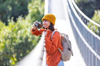 Seyahat ve fotoğrafçılık. Elinde kamera ve sırt çantası olan genç bir kadın asma köprü boyunca yürüyor. Gezinin ve fotoğraf çekmenin keyfini çıkaran mutlu bayan turist. Kameralı kız..