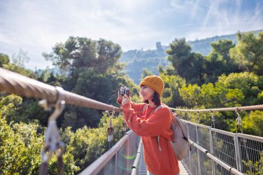 Seyahat ve fotoğrafçılık. Elinde kamera ve sırt çantası olan genç bir kadın asma köprü boyunca yürüyor. Seyahatinin tadını çıkaran mutlu bayan turist. Kameralı kız..