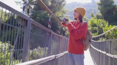 Seyahat ve fotoğrafçılık. Elinde kamera ve sırt çantası olan genç bir kadın asma köprü boyunca yürüyor. Gezinin ve fotoğraf çekmenin keyfini çıkaran mutlu bayan turist. Kameralı kız..
