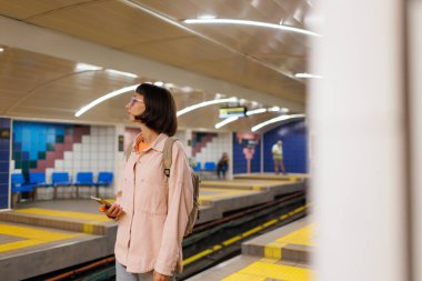 Genç bir kadın metro istasyonunda duruyor ve cep telefonuna bakıyor. Bir kadın metro istasyonunda cep telefonu kullanıyor. Kız programa bakıyor..