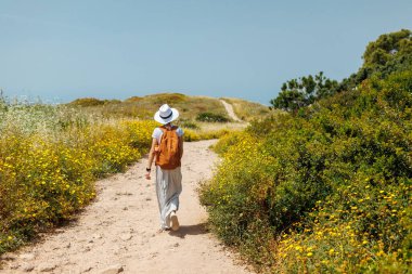 Genç bir kız sırt çantasıyla yolda yürüyor ve beyaz bir elbise giyiyor. Elbiseli ve sırt çantalı bir kız. Mutlu ve özgürlük düşkünü bir kadın yürüyüş yapıyor. Yaz havası