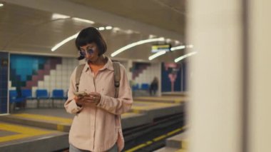 Kız, treni bekliyor ve telefonu kullanıyor. Metrodaki kız. Metroda sırt çantasıyla şık giyinmiş genç bir kız. moda ve tarz.