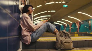 Kız, treni bekliyor ve telefonu kullanıyor. Metrodaki kız. Metroda sırt çantasıyla şık giyinmiş genç bir kız. moda ve tarz.