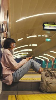 Genç kız istasyonda tren bekliyor ve telefonu kullanıyor. Uygulamada görüntüleme çizelgesi. Toplu taşıma araçlarıyla seyahat etmek. Metroda sırt çantalı şık elbiseli genç bir kız..