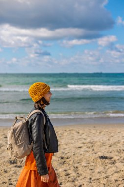 Soğuk havada kumsalda sırt çantalı bir kadın. Güneşli bir günde deniz kenarında yürüyüş yapan şık bir kadın. Sırt çantalı kız..