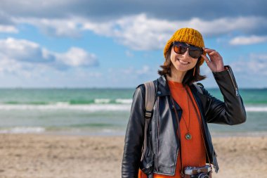 Sırt çantası ve kamerası olan bir kız. Sırt çantalı ve kameralı şık bir kadın soğuk ve güneşli bir günde plaja gitmekten hoşlanır. Yaşam tarzı, seyahat, turizm, doğa, aktif yaşam.