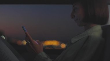 Genç bir kadın kanepede pencerenin yanında otururken telefonu kullanıyor ve gece şehrin ışıklarına bakıyor. Kız elinde telefonla dinleniyor..