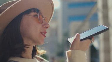 Genç ve çekici bir kadın parkta bir bankta oturuyor ve hoparlörle konuşuyor, genç nesil modern teknolojiyi kullanmaktan memnun. Sesli mesaj gönder, sesli mesaj gönder, haberleri paylaş. 