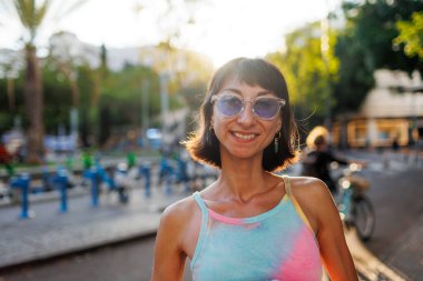 Neşeli, neşeli, genç bir kadının portresi. Hoş bir gülümsemesi var. Şık kıyafetler giyiyor ve güneşli bir günde açık havada güneş gözlüğü takıyor. Kız şehirde yürümekten zevk alıyor..