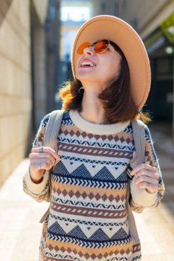 Şehirde sırt çantalı bir kız. Binaların arasında cadde boyunca yürüyen mutlu genç bir kadın. Sırt çantalı bir bayan gezgin şehri geziyor, şık bir kız şehirde dolaşıyor..