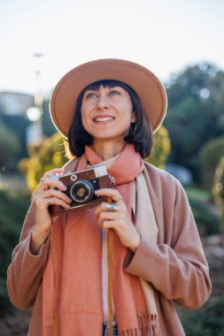 Şık bir görünüş, kameralı genç bir kadın zarif bir şapka ve ceket giyiyor. Hipster bir turist kadın elinde kamerayla fotoğraf çekiyor. film kamerası.