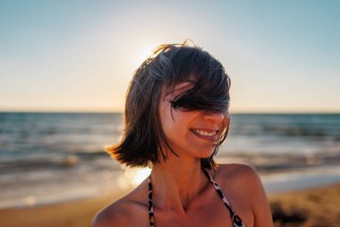 Genç mutlu kız gülümsüyor. Saçında rüzgar var. Saç yüzünü kapatır. Sahilde genç ve güzel bir kadının portresi. Deniz kıyısındaki genç kız