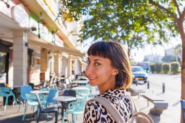 Sırt çantasıyla şehirde dolaşan bir kadın. Şehrin etrafında dolaş. Leopar desenli bluz giyen ve sırtında sırt çantasıyla bir yaz günü sokakta yürüyen neşeli bir kadın..