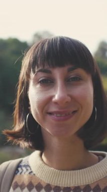 Turist kameraya bakar ve gülümser. Süslü bir kazak giyen ve sırt çantalı bir kız ormanda yürümekten zevk alıyor. Sırt çantalı bir kız ormanda yürüyor. Yürüyüş ve yürüyüş.