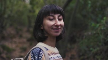 Turist kameraya bakar ve gülümser. Süslü bir kazak giyen ve sırt çantalı bir kız ormanda yürümekten zevk alıyor. Sırt çantalı bir kız ormanda yürüyor. Yürüyüş ve yürüyüş.
