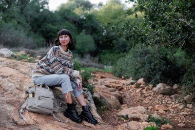 Sırt çantalı ve süveterli bir yürüyüşçü ormanda bir taşın üzerinde oturuyor.