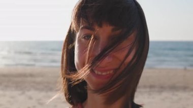 Yaz havası. Kumsalda güzel bir esmerin portresi. Genç mutlu kız gülümsüyor. Saçında rüzgar var. Saç yüzünü kapatır. Genç bir kız sahilde eğleniyor..