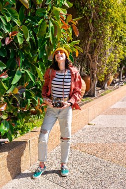 Şehir caddesinde kameralı ve sırt çantalı mutlu genç kız. Şehirde dolaşan bir turist. Sokak bitkileri. Şehrin arka planında neşeli, güzel turistin açık hava portresi.