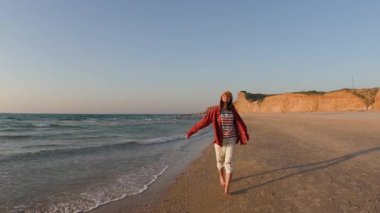 Sahilde yürüyen gömlekli bir kadın ve arkasında güzel bir günbatımı var. Yaz tatilindeki turist. Gülümseyen neşeli bayan sahilde eğleniyor ve güzel doğanın tadını çıkarıyor.. 